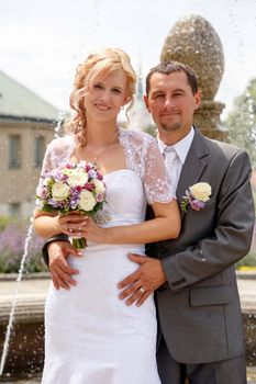 beautiful young wedding couple, blonde bride with flower and her groom against fountain