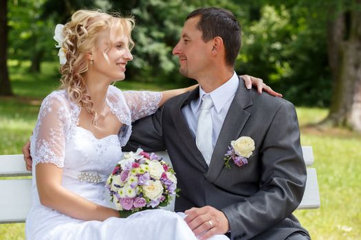 beautiful young wedding couple, blonde bride with flower and her groom looking to each other on park bench