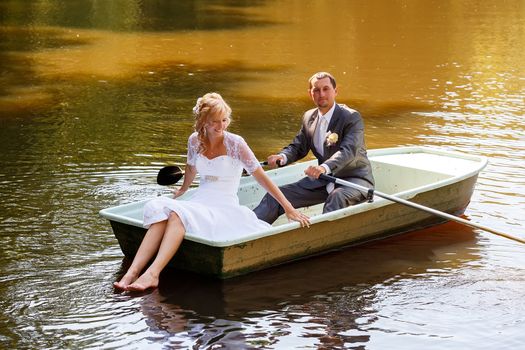 beautiful young wedding couple, blonde bride with flower and her groom just married on small boat at pond with evening sun