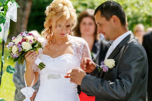 beautiful young couple in wedding ceremony outdoor, blonde bride with flower and groom exchange of wedding rings