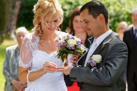 beautiful young couple in wedding ceremony outdoor, blonde bride with flower and groom exchange of wedding rings