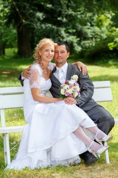 beautiful young wedding couple in park, sitting on bench, blonde bride with flower and her groom