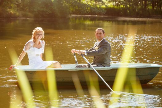 beautiful young wedding couple, blonde bride with flower and her groom just married on small boat at pond with evening sun