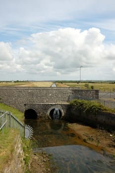 A river flows from marshland through a pair of tunnels under a stone bridge.