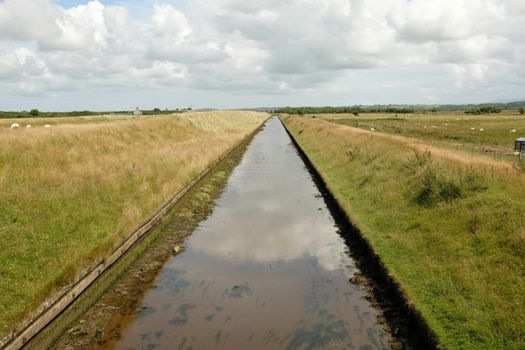 A constructed tidal canal with high grass banks leads through marshland to the horizon with a cloudy sky.