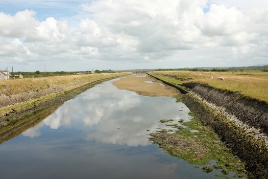 A constructed tidal river with high grass banks leads through marshland to the horizon with a cloudy sky.