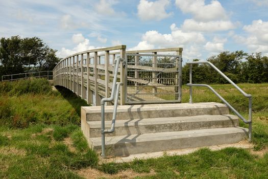 Concrete steps lead to a gate and foot bridge with wooden rails with a blue cloudy sky in the background.