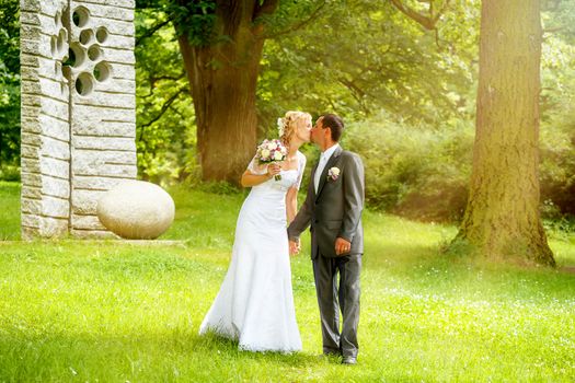 beautiful young wedding couple kissing , blonde bride with flower and her groom in summer park with sun haze