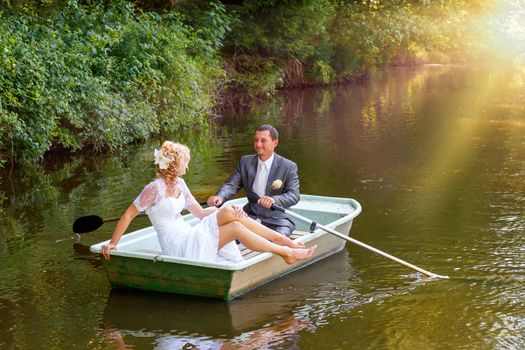 beautiful young wedding couple, blonde bride with flower and her groom just married on small boat at pond with evening sun