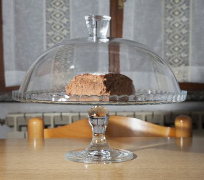 Chocolate cake inside a glass stand, over kitchen table