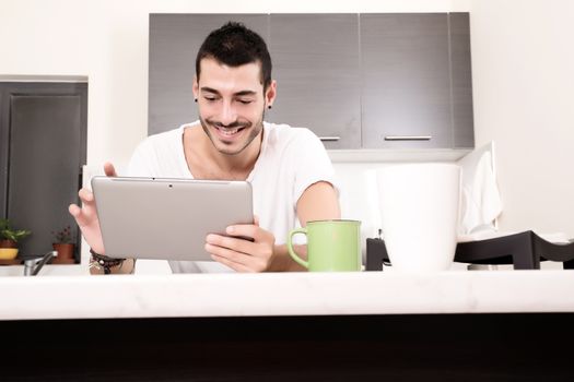 A young male sitting in the kitchen with a Tablet PC.