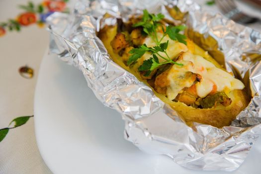 Baked potato with vegetables,chicken filet,fresh parsley and cheese in the aluminium foil  on the white plate