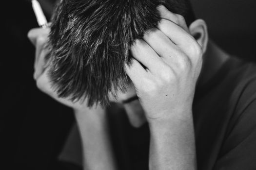 Black and white portrait of a teenage boy leaning his head against his fists while holding a pen, expressing frustration, negative feeling or intellectual effort, on black