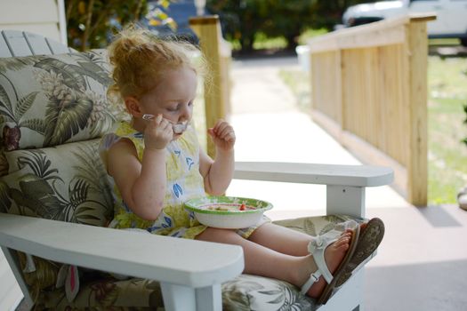 Cute little girl with summer dress eating by herself a nutritious snack in a bowl while sitting on an armchair with floral pattern, outdoors, on the porch