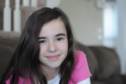 Portrait of a brunette long-haired pretty teenage girl smiling at camera with a happy and confident facial expression, wearing a pink T-shirt, indoors