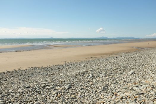 Pebbles lead down to a deserted sandy beach and sea with mountains in the distance.