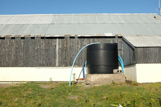 A black plastic water storage tank with blue pipes against a wooden building with tin roof.