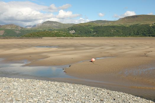 Pebbles lead down to a deserted sandy estuary with a buoy at low tide, the mountain Cadair Idris seen in the distance, Gwynedd, Wales, UK.