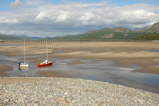 A person preparing sail boats on the sand of an estuary at low tide with mountains in the distance.