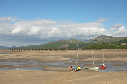 People preparing sail boats on the sand of an estuary at low tide with mountains in the distance.