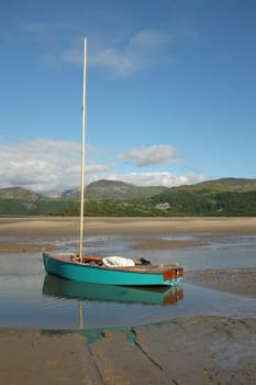 A turquoise sail dinghy floats in a puddle on tha sand of an estuary with mountains in the distance.