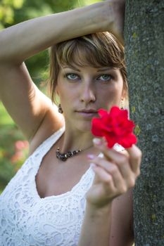 Attractive woman in a stylish white lacy top holding out a fragrant red rose symbolic of love as she leans against a tree in the garden
