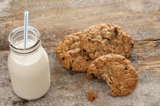 Small glass bottle of fresh creamy farm milk with a straw alongside half eaten crunchy cookies on a rustic wooden surface, high angle view