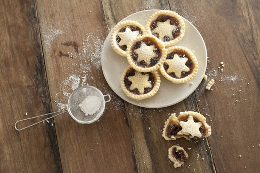 Overhead view of a plate of decorative freshly baked Christmas mince pies with pastry stars alongside a half eaten pie and strainer with icing sugar to dust the top