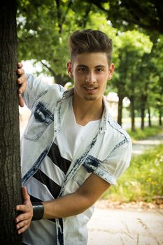 Friendly handsome young man smiling at camera, behind tree
