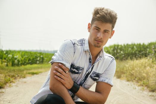 Handsome young man sitting in the middle of coutryside dirt road, looking at camera