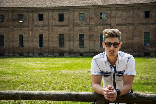 Handsome young man outdoors leaning on wood fence, old house or farm behind him