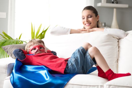 Mother looking at little superhero lying down on sofa in the living room.