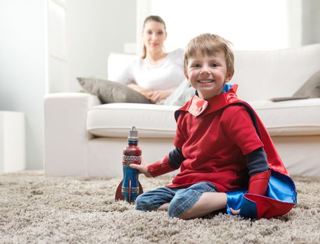 Cute superhero boy paying with toy rocket in the living room with his mother on background.