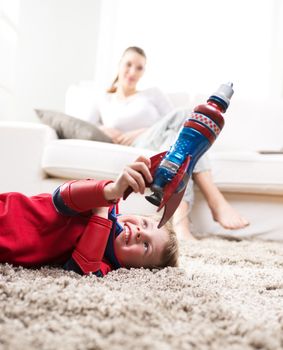 Cute superhero boy paying with toy rocket in the living room with his mother on background.