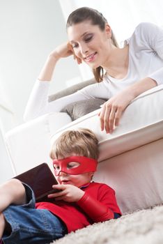 Cute superhero boy playing videogames sitting on carpet with his mother on background.