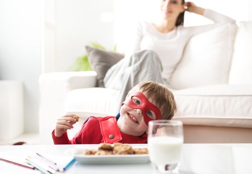 Superhero boy having an healthy snack with cookies and milk with his mother on background.