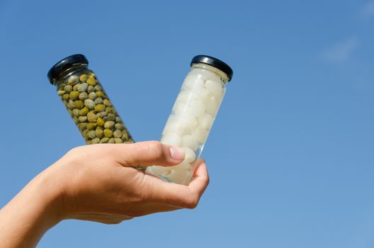 hand hold two marinated capers and garlic glass jar on blue sky background