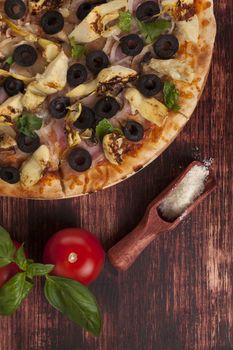 Rustic, country style pizza background. Pizza with black olives, ham and artichokes on wooden background with parmigiano cheese, tomatoes and basil.