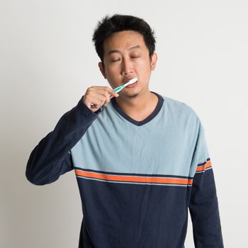 Sleepy Southeast Asian male brushing teeth while eyes closing in a morning, on plain background