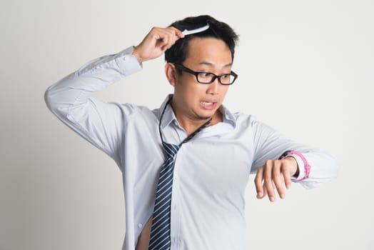Asian businessman combing hair in morning in hurry, checking time on watch, on plain background.