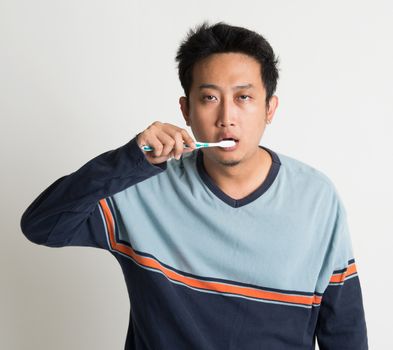Southeast Asian male brushing teeth with sleepy eyes in a morning, on plain background