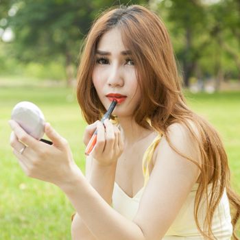 Woman applying lipstick. Asian woman with long hair sitting on the grass in the park. In applying lipstick