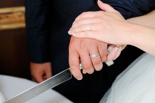 Closeup of bride and groom cutting wedding cake at reception showing rings and knife