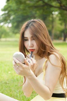 Woman applying lipstick. Asian woman with long hair sitting on the grass in the park. In applying lipstick