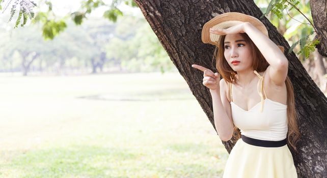 Portrait Asian woman under a tree. Woman with long hair wearing a hat is pointing forward.
