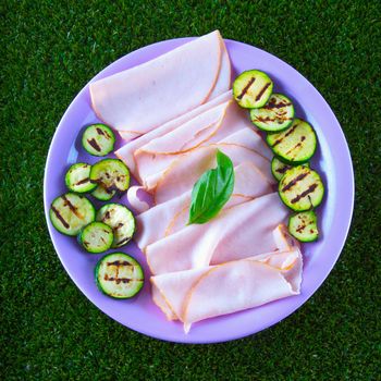 Turkey ham with grilled zucchini in a purple plate, over grass