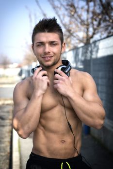 Muscular shirtless young man outdoors with headphones smiling and looking at camera
