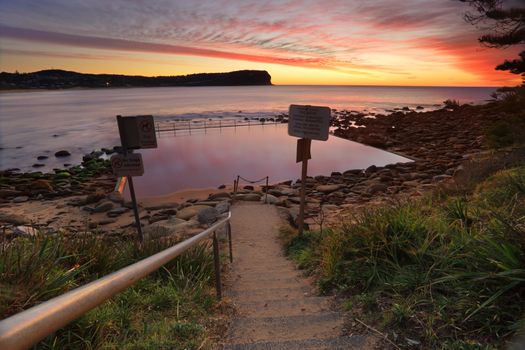 Low tide exposes the rocky end of Macmasters Beach where the tidal pool is calm reflecting pretty patterns in the sunrise sky