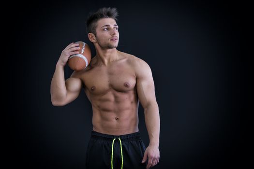 Muscular american football player standing with ball in hand, on dark background