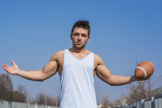 Muscular american football player standing with ball in hand, arms open. Outdoors shot
